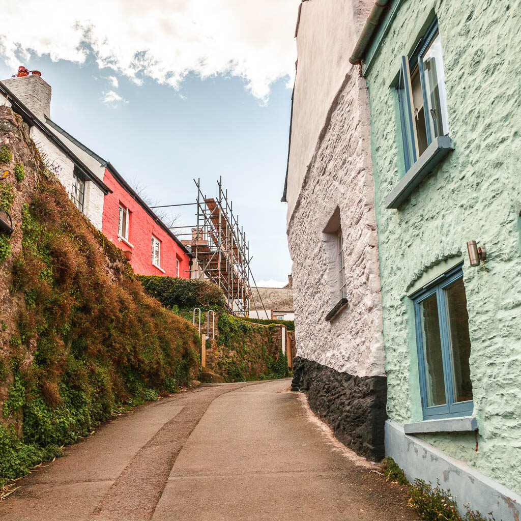 A hedge on the left of the road and turquoise and white coloured stone buildings to the right on the walk from Dittisham to Dartmouth.