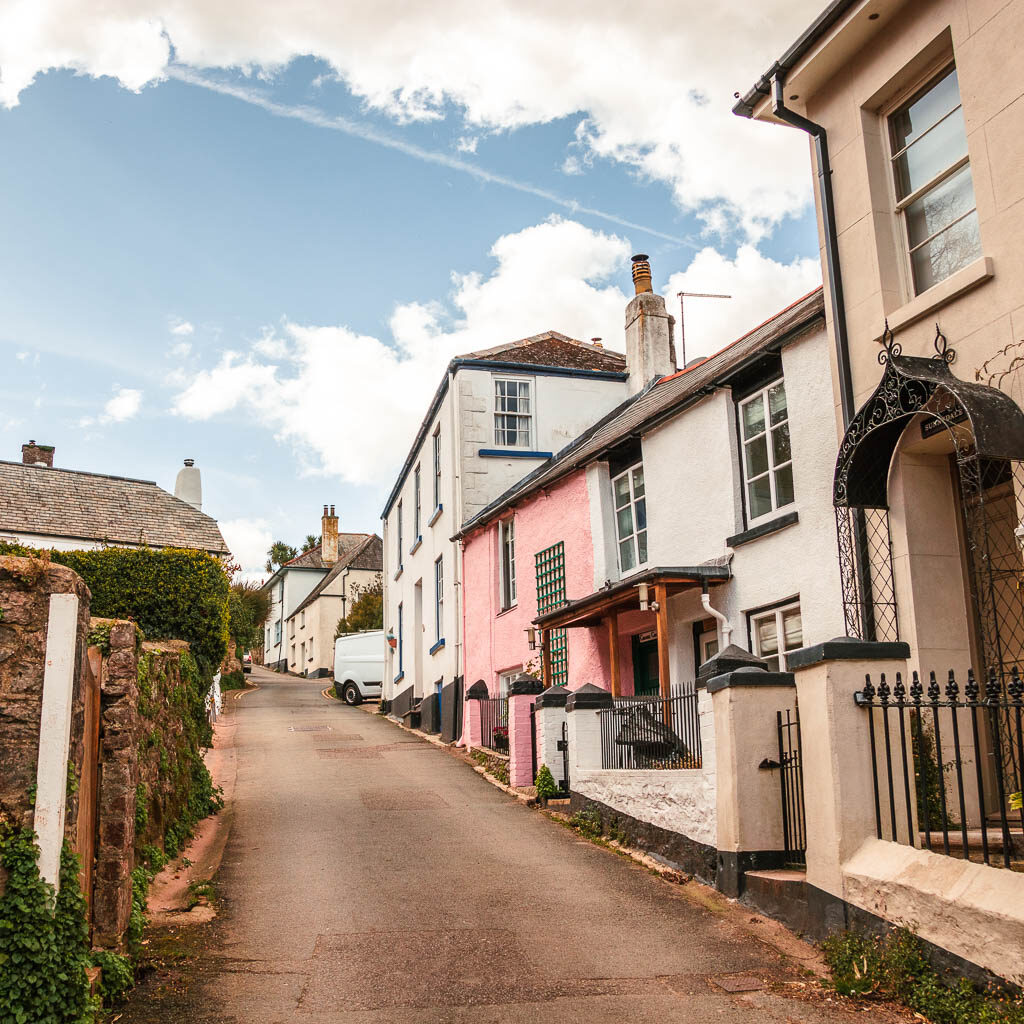 A road leading uphill, with pastel coloured houses of Dittisham to the right, on the walk to Dartmouth.