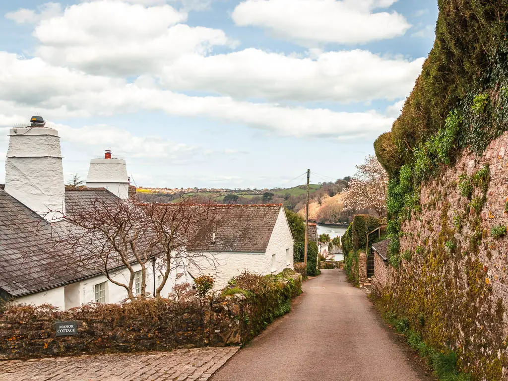 A road leading downhill, with a stone wall to the right, and white coloured stone buildings to the left on the Dartmouth Dittisham walk. Right at the bottom of the road you can see a peak of the river.