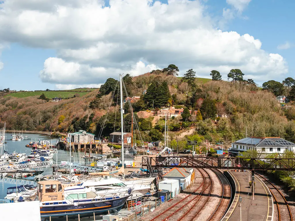 Looking down at the railway track as it curves to the left around the river Dart on the walk from Kingswear to Greenway. There are lots of boats moored on the side of the river and lots of trees on a hill ahead.