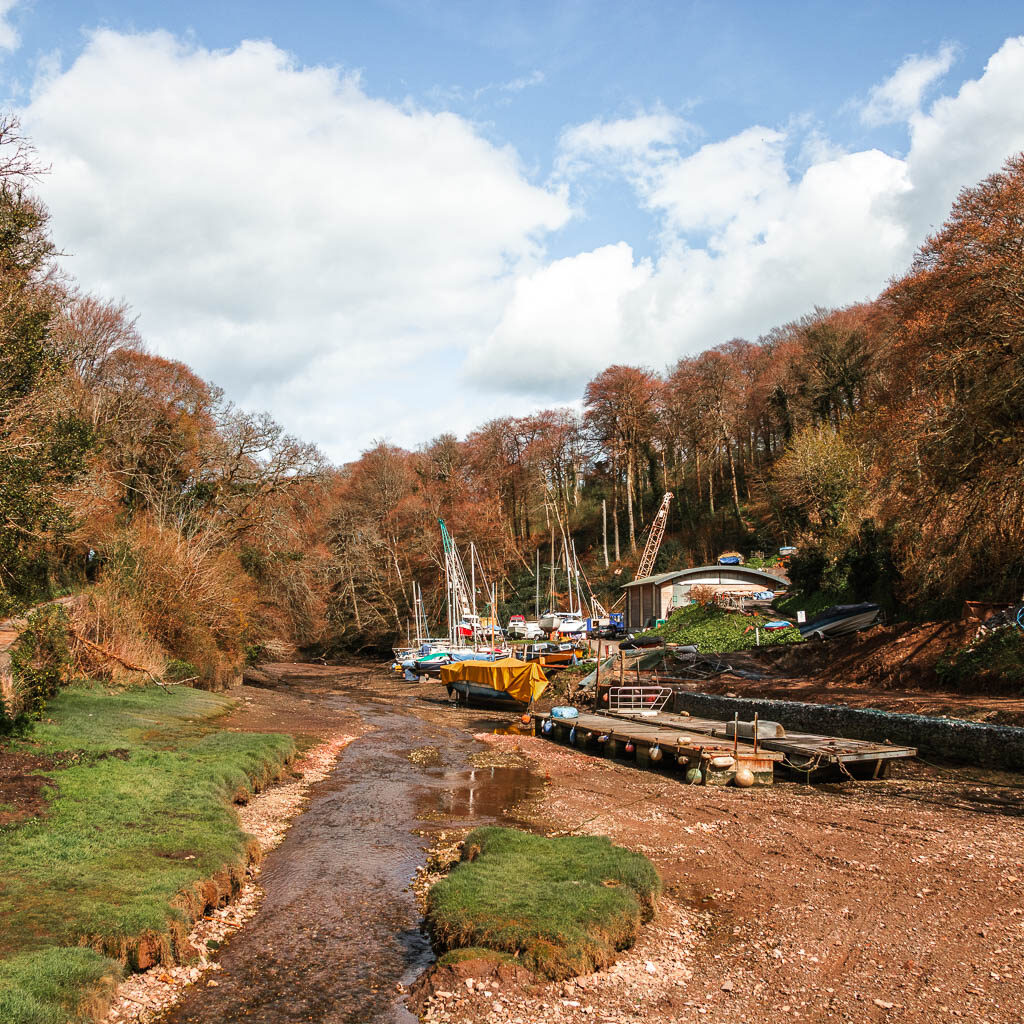 Looking along the river bed with a. few boats. There is woodland trees on both sides of the river.