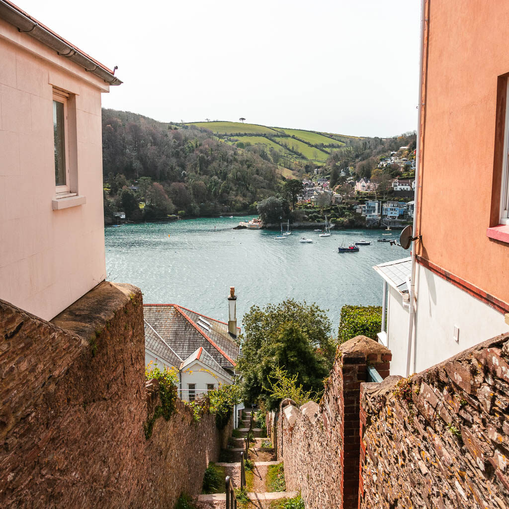 Looking down through a gap in the buildings towards the rover dar at the start of the Kingswear to Brixham coastal walk. There is a big green grass covered hill on the other side of the river.