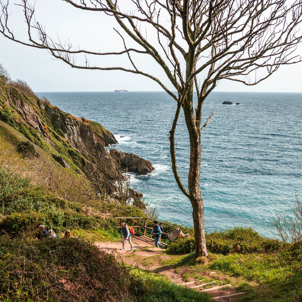 Looking down the hill to the cliff on the left and sea on the right. There are a couple of people walking on the trail below.