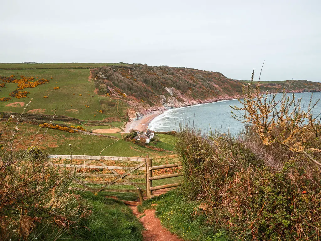 A wooden fence and gate leading down into the cliff valley with a beach and white cottage visible below.