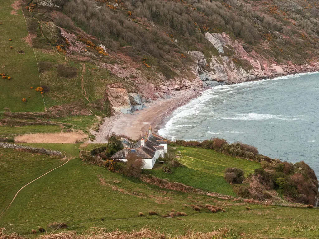 Looking down the grass hill to a white cottage in the valley on the Kingswear to Brixham coastal walk. There is a stretch of beach on the other side of the cottage and the hill and cliff on the other side. 