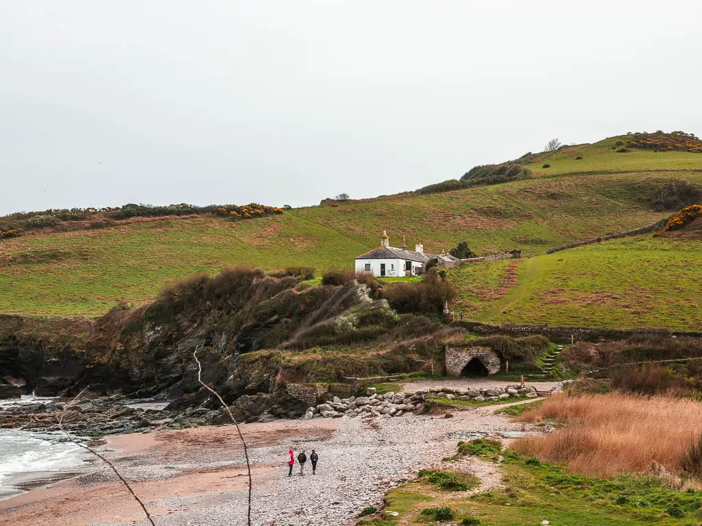 Looking across the beach to a white cottage on the cliffside on the Kingswear to Brixham coastal walk. There are a few people walking on the beach.