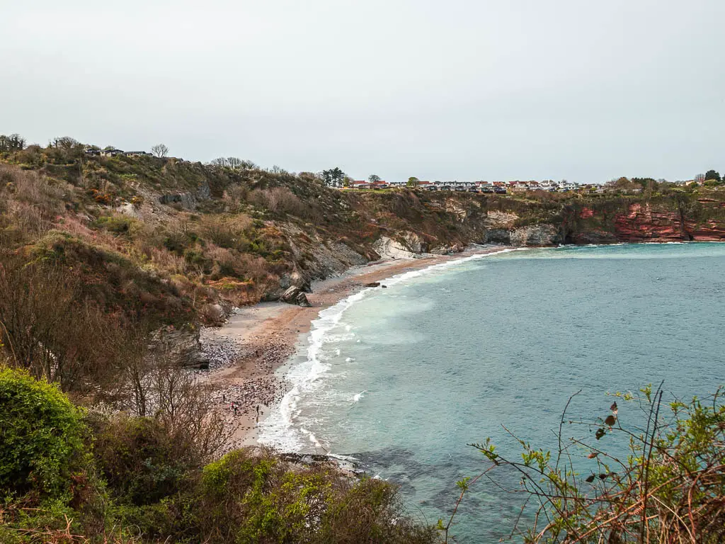 Looking down at the long stretch of sand of the beach cove as it curves around. Behind it is a tree covered hill.