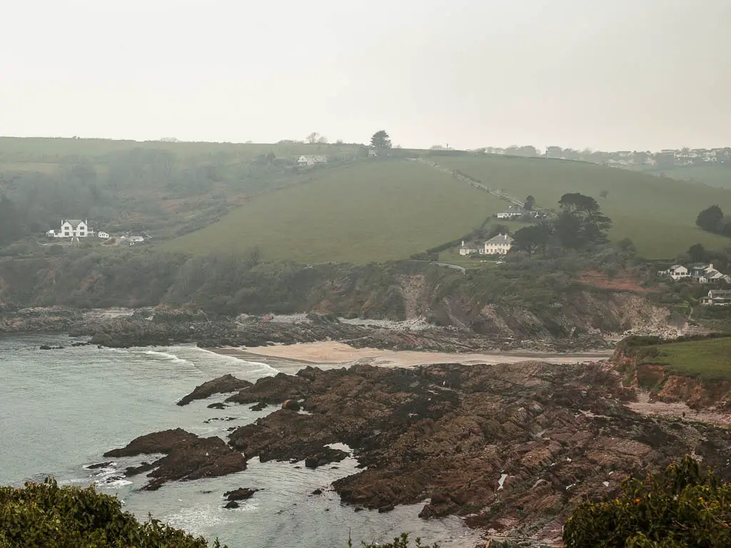 Looking down the Talland bay, part way through the walk from Looe to Polperro. Its a cove with a small beach and lots of rocks. There are a few houses on the cliff hill on the other side of the cove. It's a foggy day.