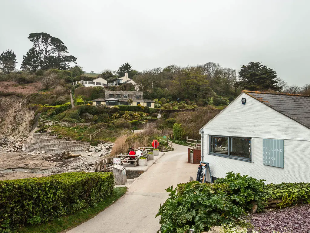 A light coloured road leading down, with the white building of the Talland Bay beach cafe on the right, and some houses on the hill ahead,