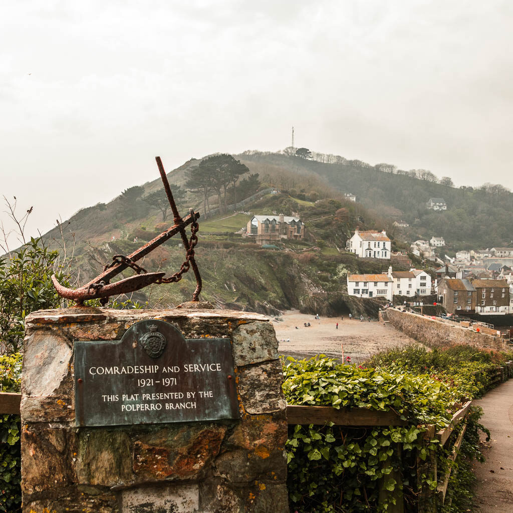 A memorial plaque and anchor pushed on top, with Poplerro beach down bellow on the other side, and the houses of Polperro to the right.