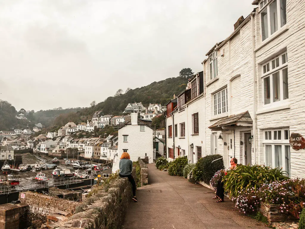 The path leading to Looe, with some white coloured buildings on the right and the harbour to the left. There is a stone wall on the left with a person sitting on it.