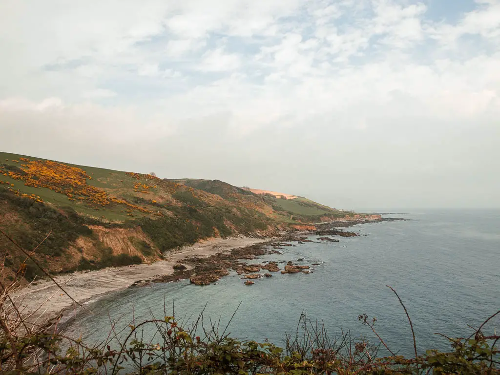 Looking across the blue sea to the grey sand and cliffs with a grass and gorse covered clifftop.