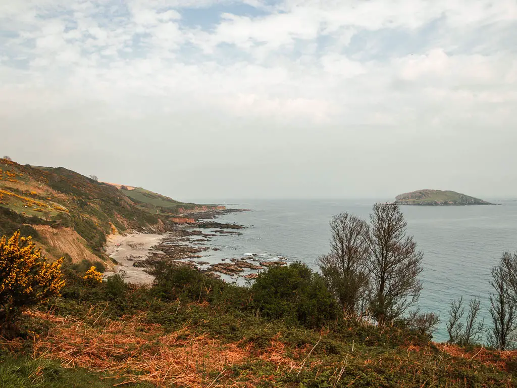 Looking along the cliffy coastline on the left, with a sandy beach in front which lead to some rocks meeting the sea. Looe Island is out at sea to the right.