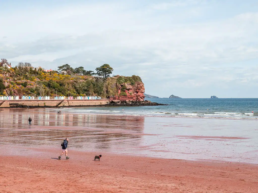 Looking across the red sand beach towards the red cliffs and beach huts on the left and sea ahead on the coastal walk from Paignton to Brixham