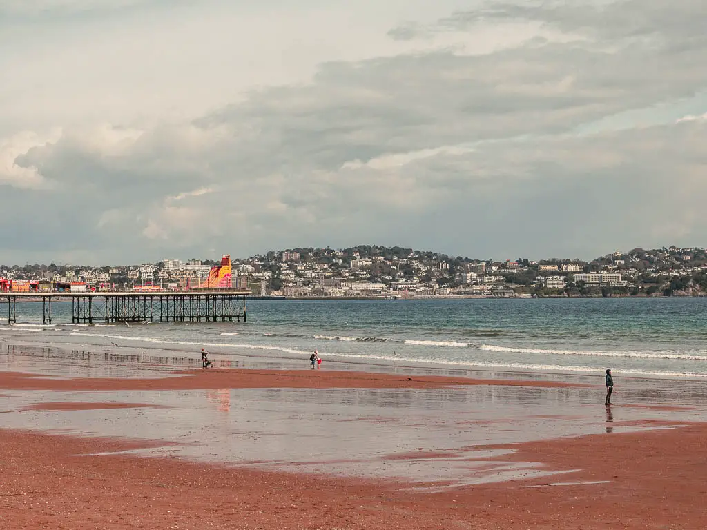 The red sand beach in Paignton at the start of the coastal walk to Brixham. The pier is jutting out to the water on the left and a hilly landscape on the other side of the water with lots of houses and buildings.