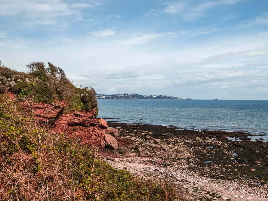 Looking down to a pebble beach cove with red cliffs on the left and the blue sea ahead.