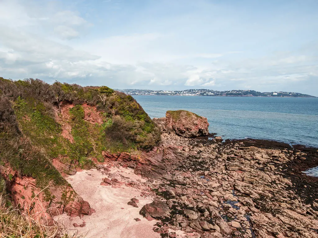 A sandy Bach cove enclosed by moss covered red cliffs on the left and the blue coloured sea ahead and right.