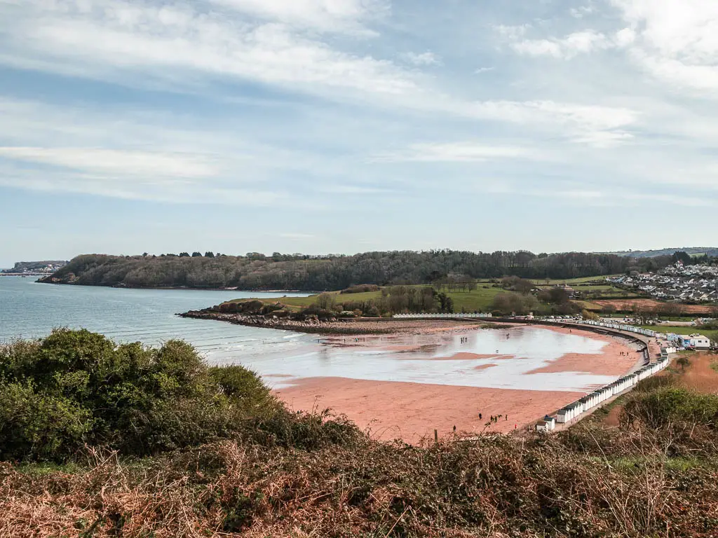 Looking down to the red sand of Broadsands Beach on the coastal walk from Paignton to Brixham. The beach is surrounded by grass and bushes. There are a few people visible walking on the beach.