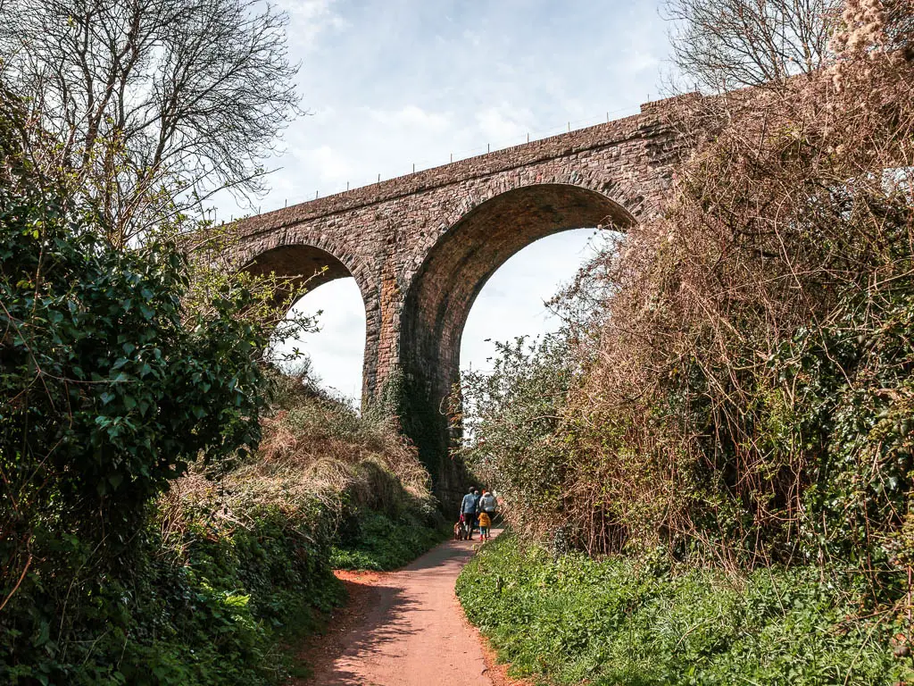 Looking under the arch of a viaduct which is surrounded by bushes on the coastal walk from Paignton to Brixham. there is a family with two adults and two children walking under the archway.