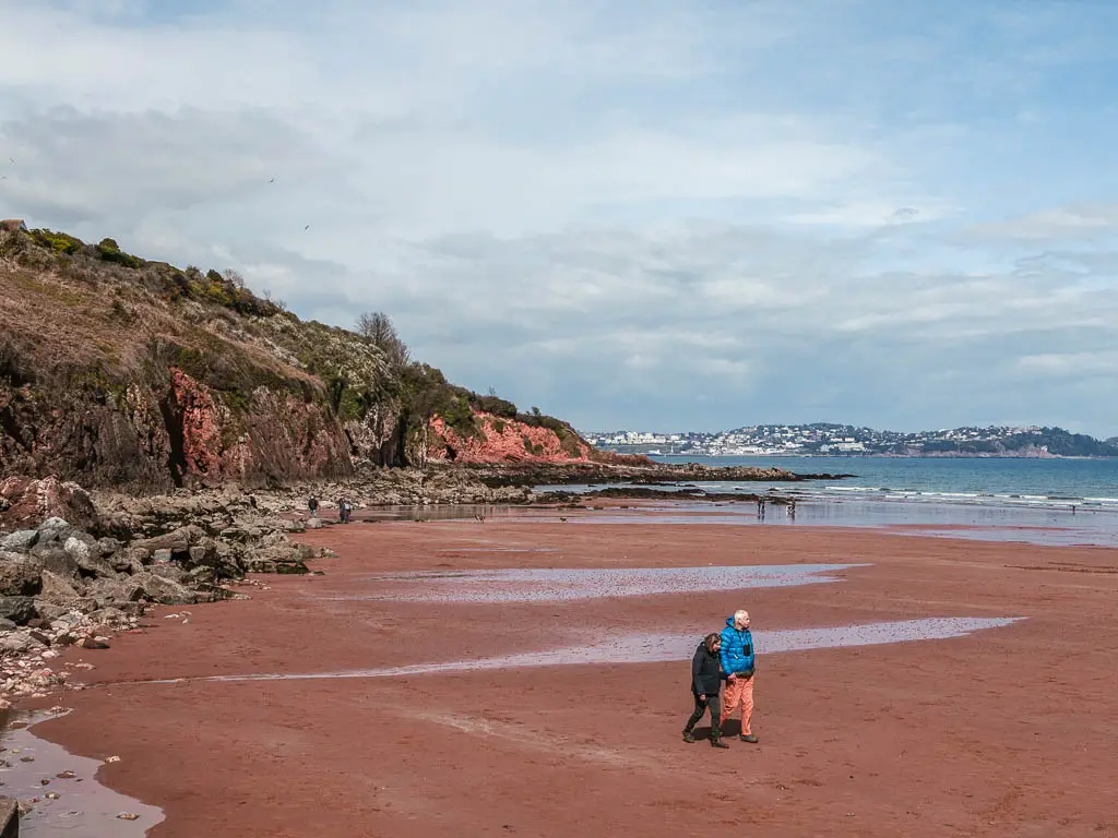 A red sand beach with cliffs on the left on the walk to Brixham from Paignton. There are a couple of people walking on the sand.