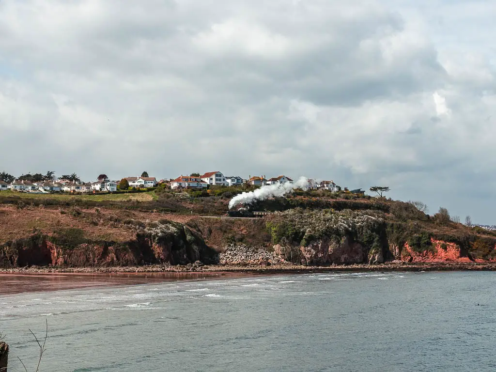 Looking across the sea with the cliffs and a steam train running across the top of them on the coastal walk from Paignton to Brixham. There are also some white coloured houses on the cliff top.