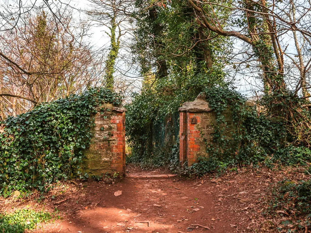 the wide dirt trail leading though short brick pillars and surrounded by trees.