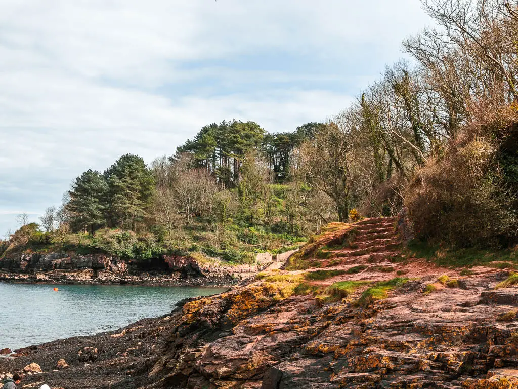 Rocky ground leading up to some rocks steps, with the sea ahead and to the left. There are trees on the peninsular on the other side of the blue water.