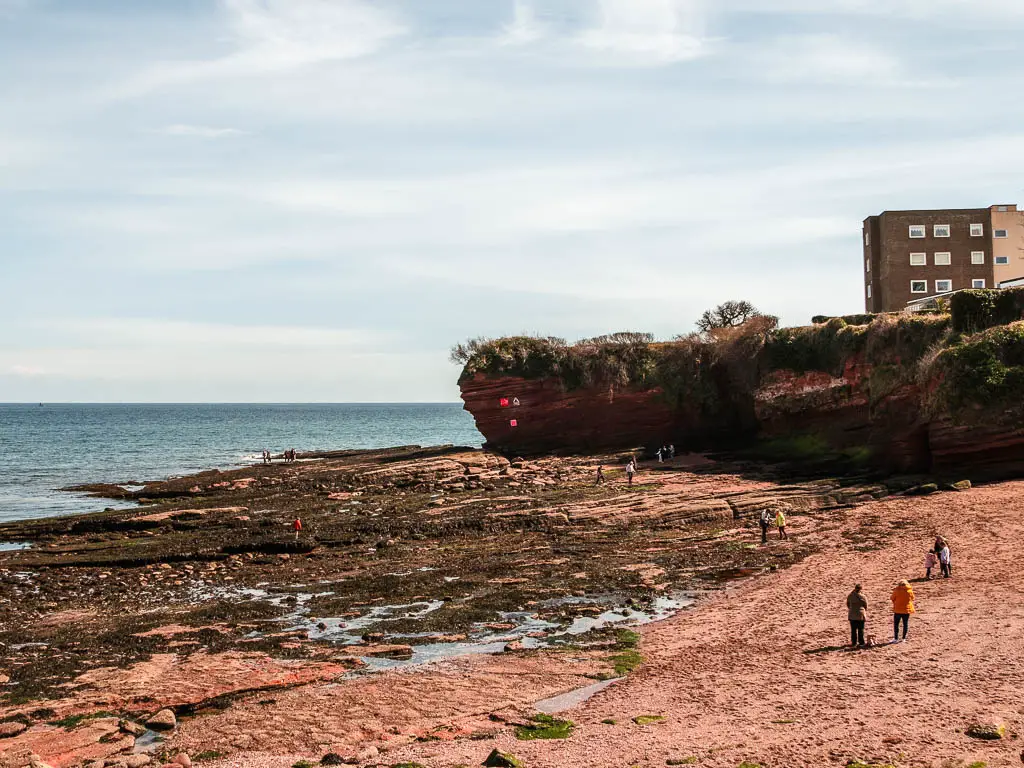 Looking down to the sandy beach with some short red cliffs ahead and the blue to to the left. There are a few people walking on the beach.