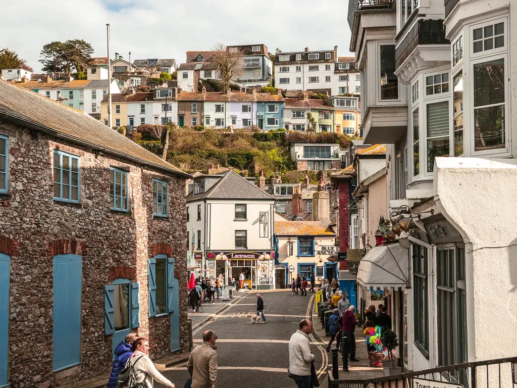 Looking down the street in Brixham and colourful buildings ahead and up the hills. There are lots of people walking about on the street.