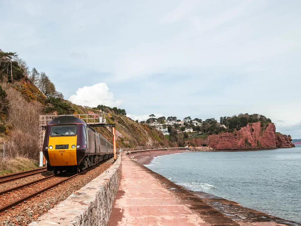 A train on the left, red cliffs ahead and the sea to the right on the Teignmouth to Dawlish railway walk.