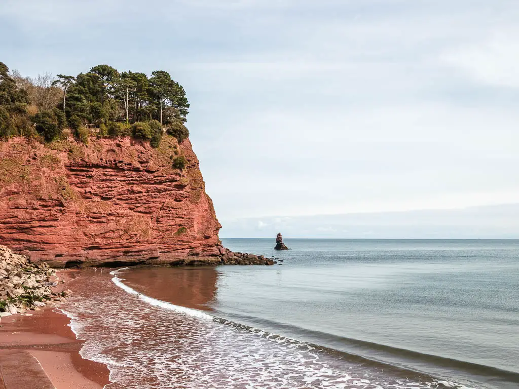 A big red cliff face ahead next to the sand beach and sea on the Teignmouth to Dawlish railway walk. There are trees on top of the rock.
