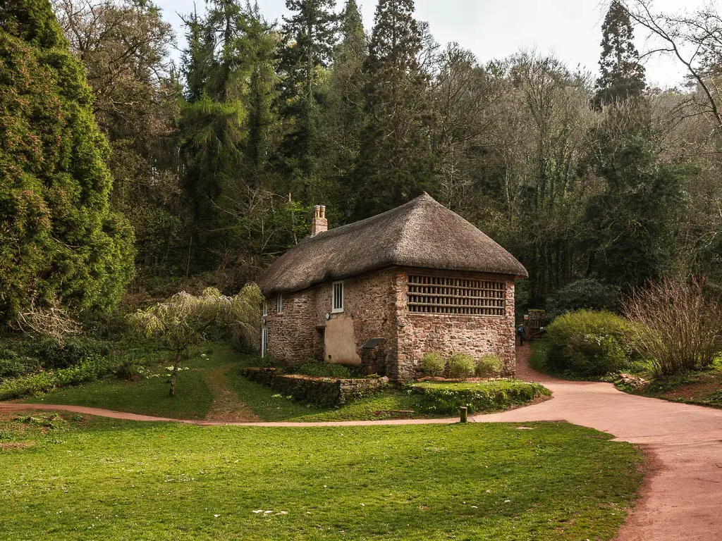 A thatched roofed cottage with stone walls on the walk from Torquay to Cockington. There is neatly cut grass in front of the cottage and woodland trees behind.