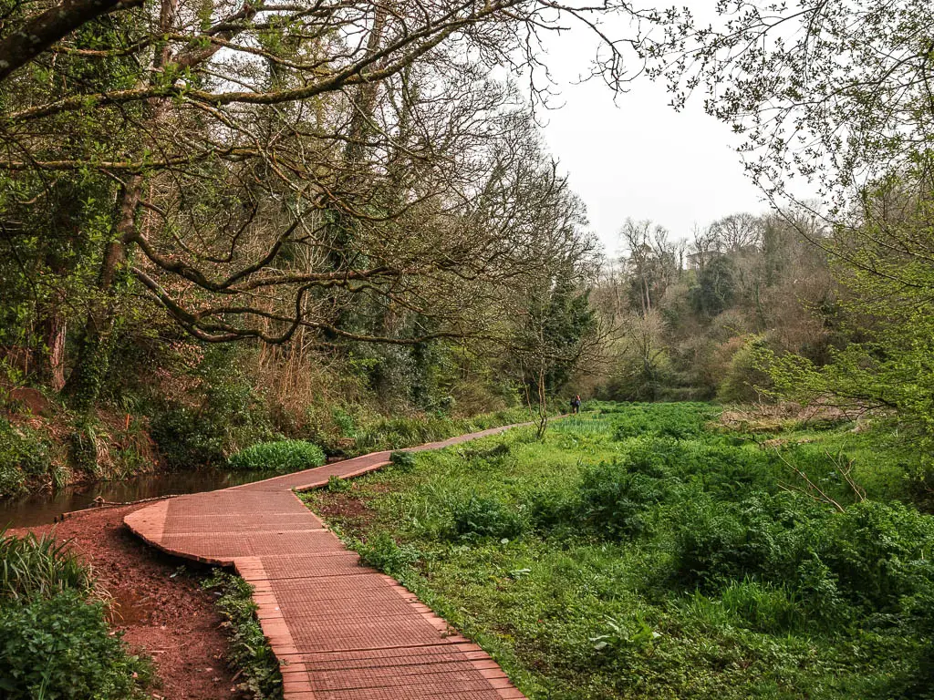 A wooden walkway with a large area of grass to the right and trees to the left, on the walk from Torquay to Cockington Village.