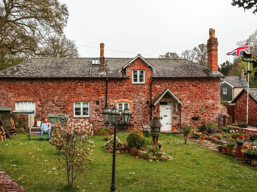 A stones walled cottage, with a front lawn with ornaments and plants in pots in Cockington. There is a Union Jack flag on the right/