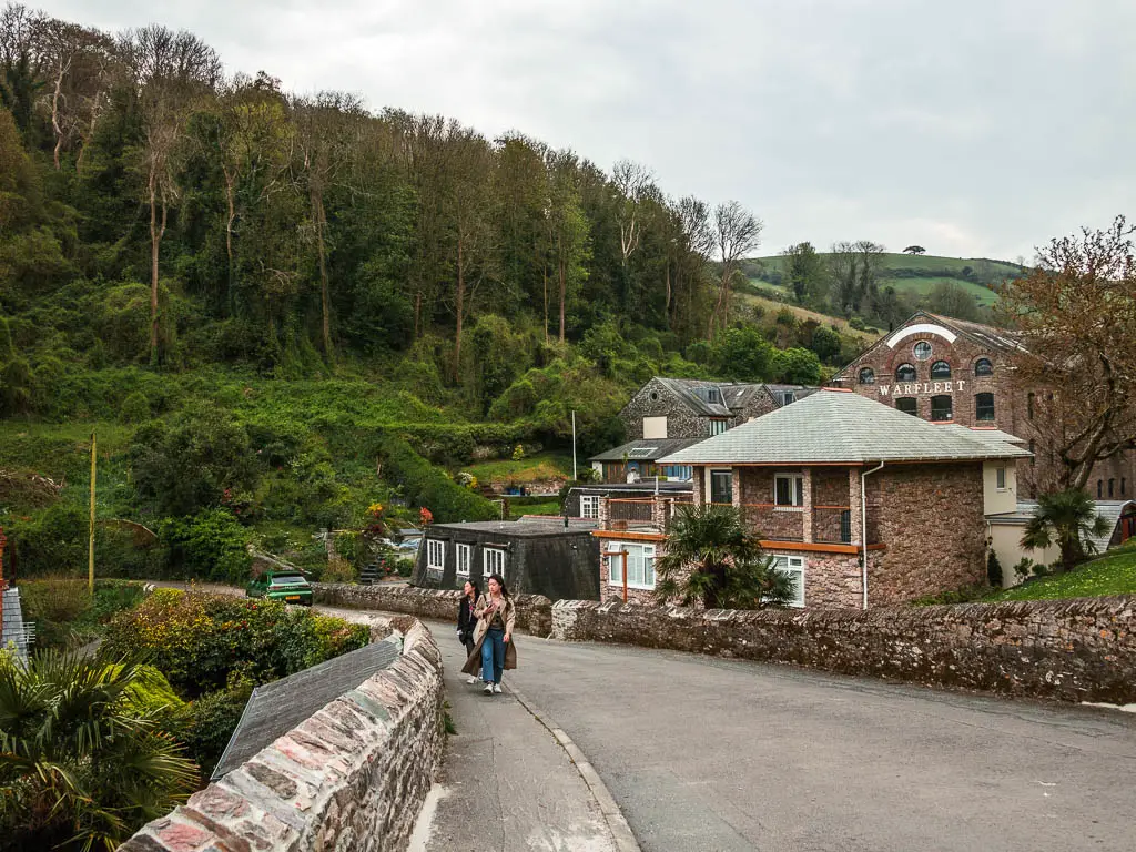 A road curving ahead to the left, lined with a stone wall on each side and some houses on the right. There are two people walking on the pavement next to the road. 