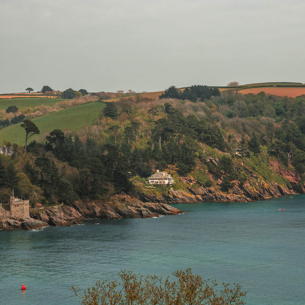 Looking across the blue sea to the cliffs on the other side along the Dartmouth to Blackpool Sands walk. There are trees along the cliff top and edge, and a houses nestled within them. 