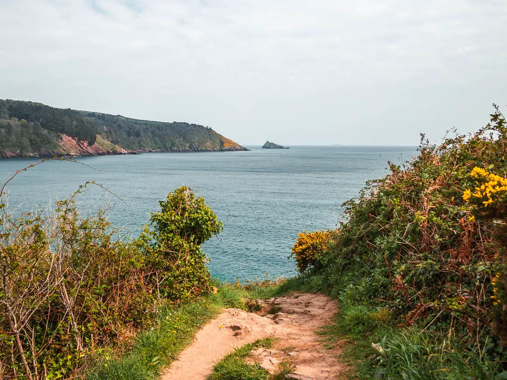 A rocky trail leading downhill, lined with some bushes, and the blue sea ahead. There is a view to a peninsular and sea rock in the distance. 