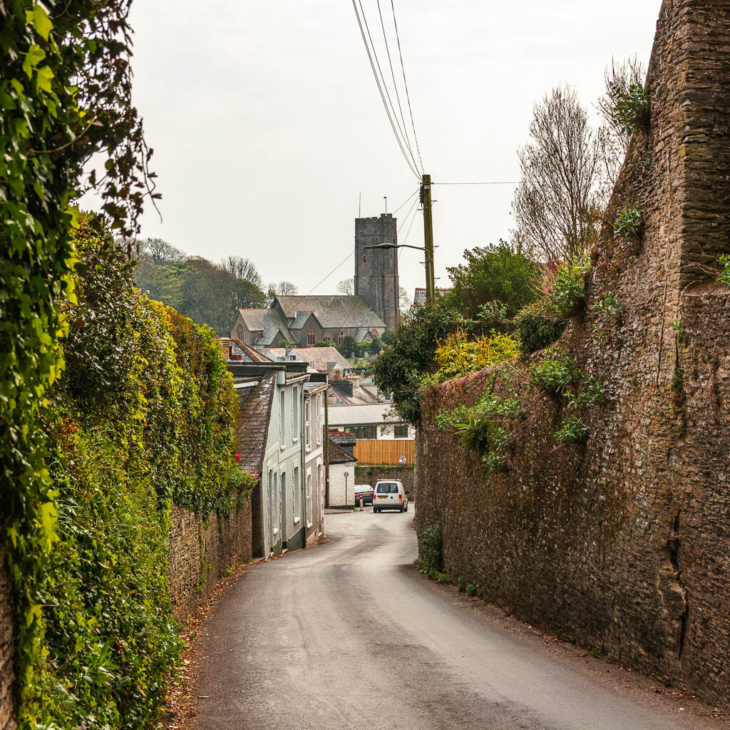 A road lined with stone walls and hedges, with some buildings of the village of Stoke Flemming ahead.