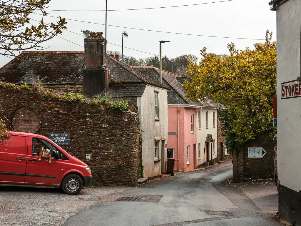 A road with some pastel coloured houses ahead on the left, and a red van with a dog hanging out the window.