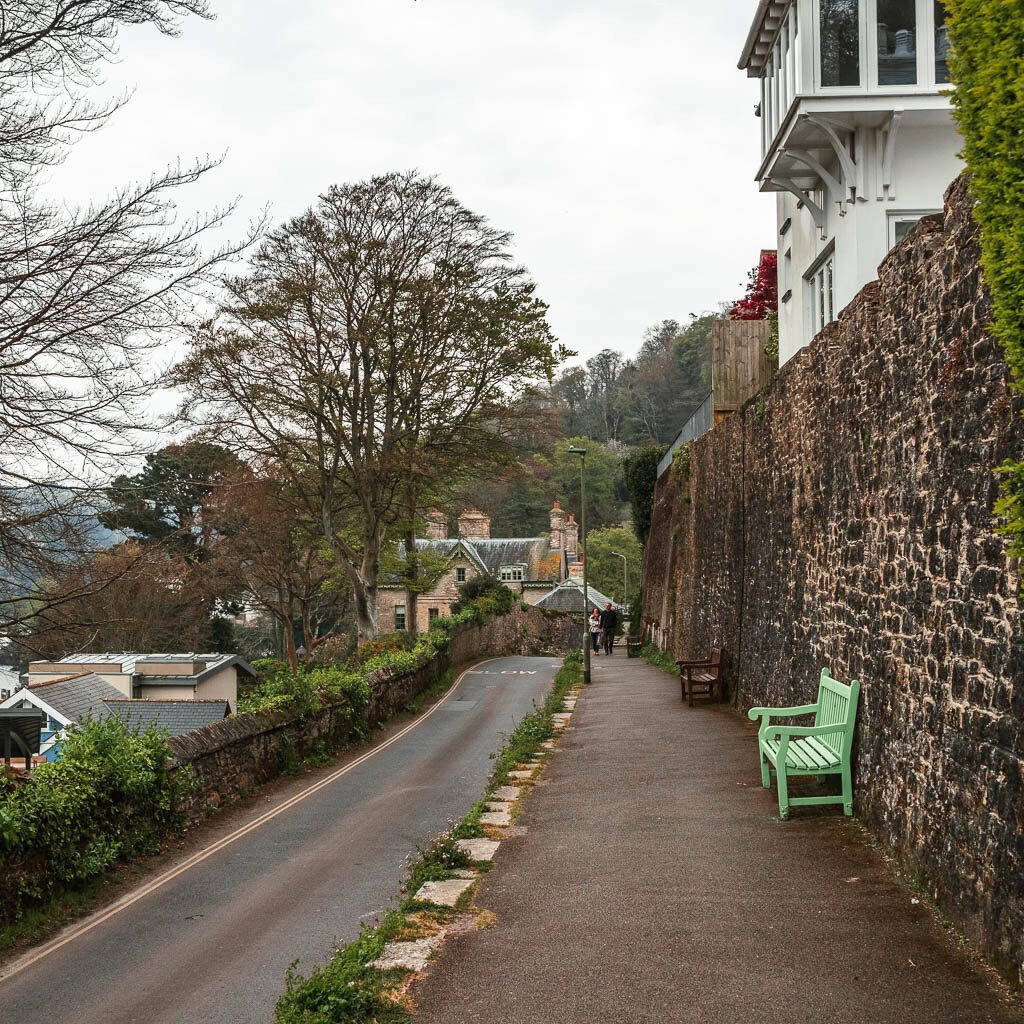 A road on the left and a raised pavement on the right. There is a stone wall to the right of the pavement, and a green bench. 