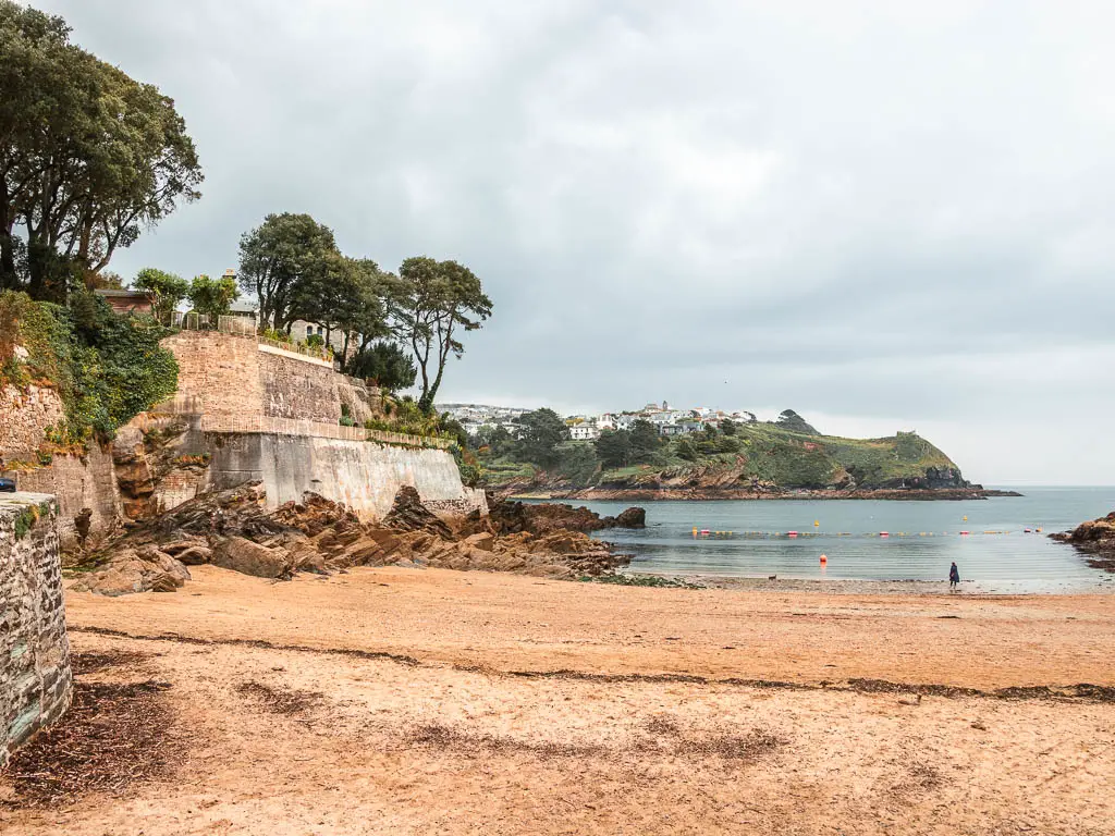 A sandy beach with stone walls on the left and the sea ahead in Fowey, at the end of the walk from Par. Polruan peninsular is across the other side of the sea. 