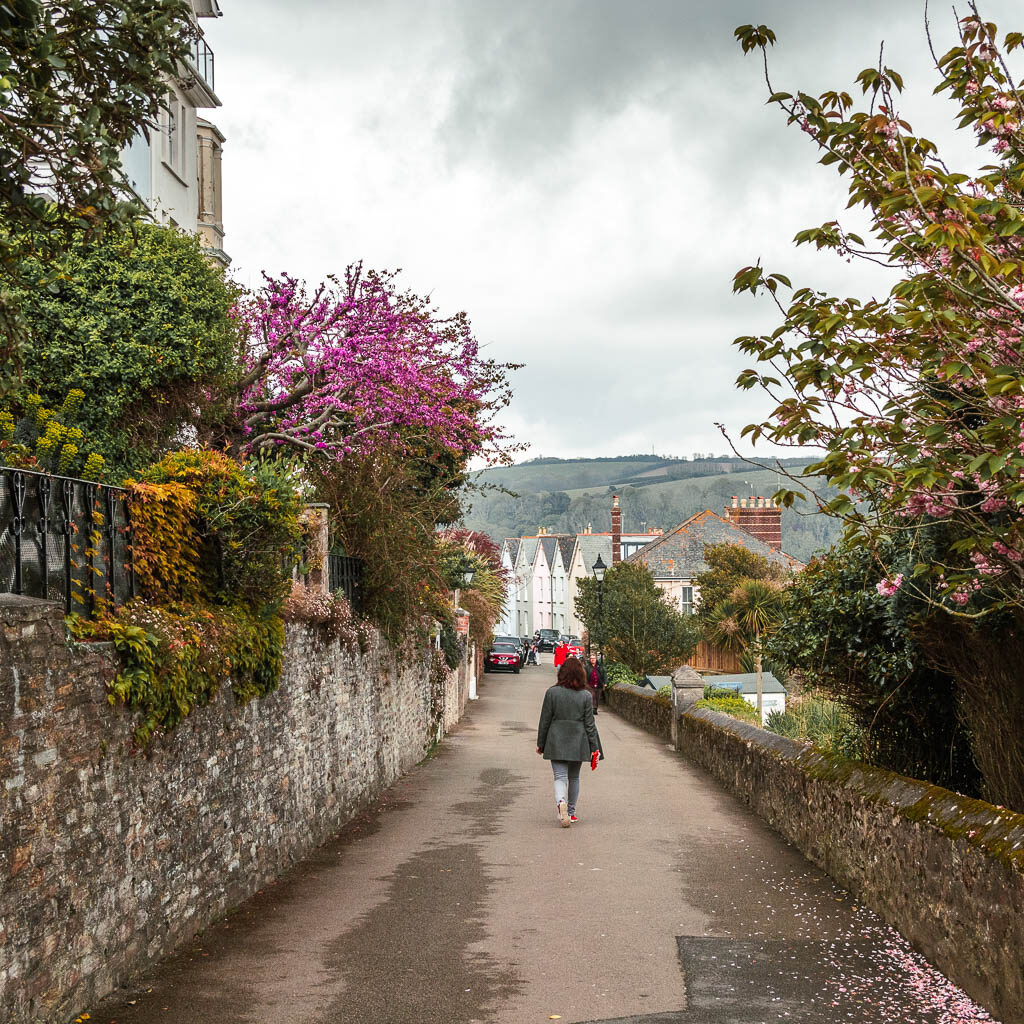 A women walking down the road in Fowey, with a tall stone wall and houses on the left, and a short stone wall on the right/