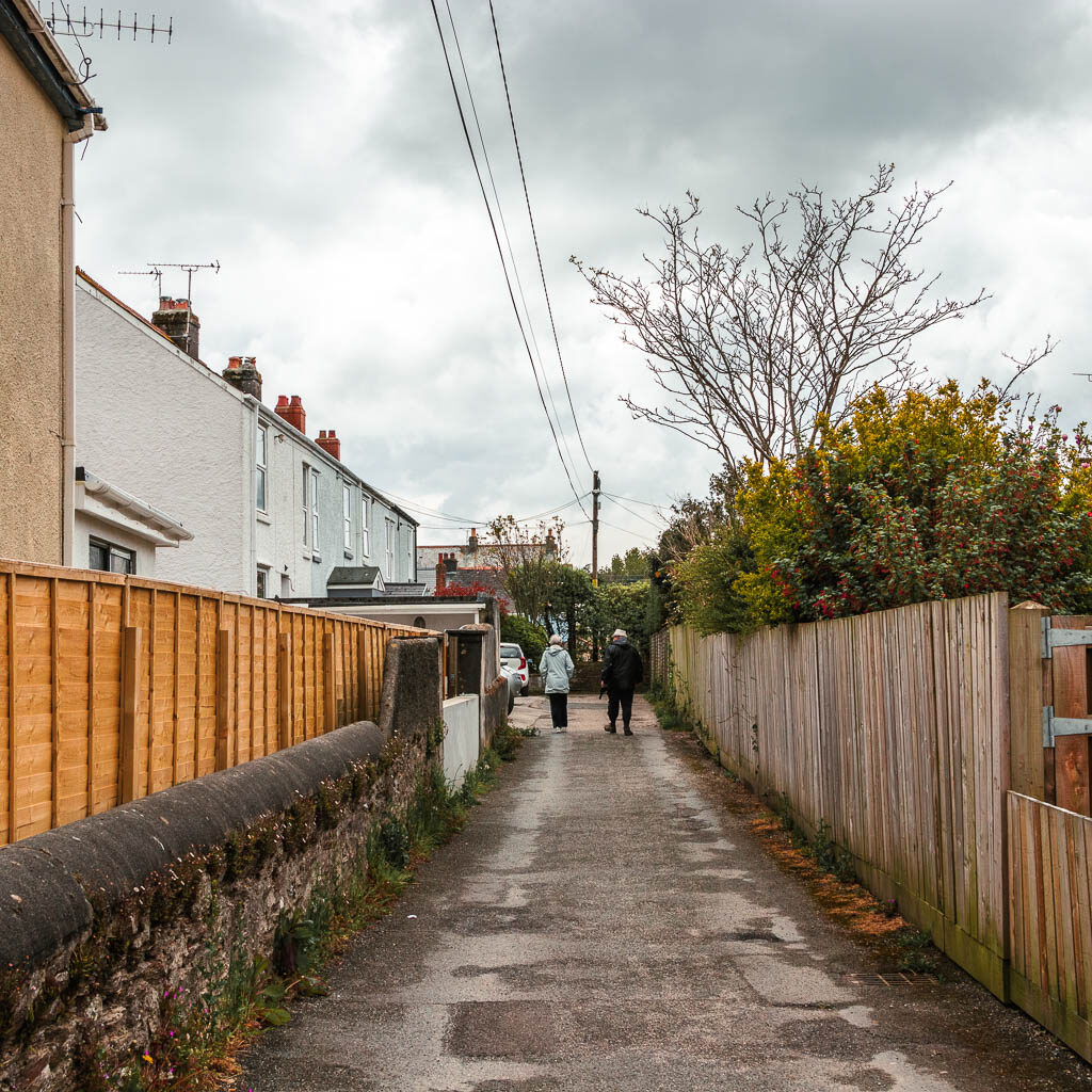 An alley walkway with a wooden fence on the right, and a wall, fence and houses on the left. There are a couple of people walking ahead on the path.