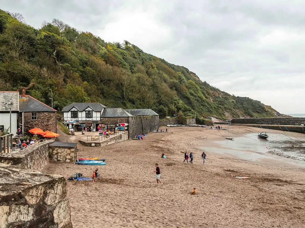 Polkaris Beach with a stone wall on the left and some tudor style buildings ahead on the walk from Par to Fowey. Behind the buildings is the tree covered cliff hill. There a re a few people walking on the beach.