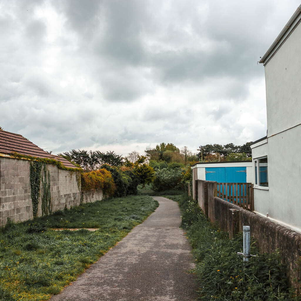 A path with a short wall and the white wall of a house on the right, and a stone wall and shed roof visible on the left.