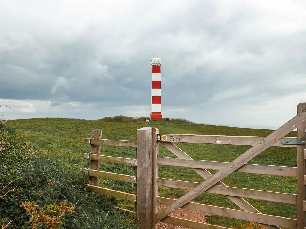 Looking past a wooden gate to the grass field and the red and white stripped Gribbin Head Daymark, on the coastal walk from Par to Fowey.