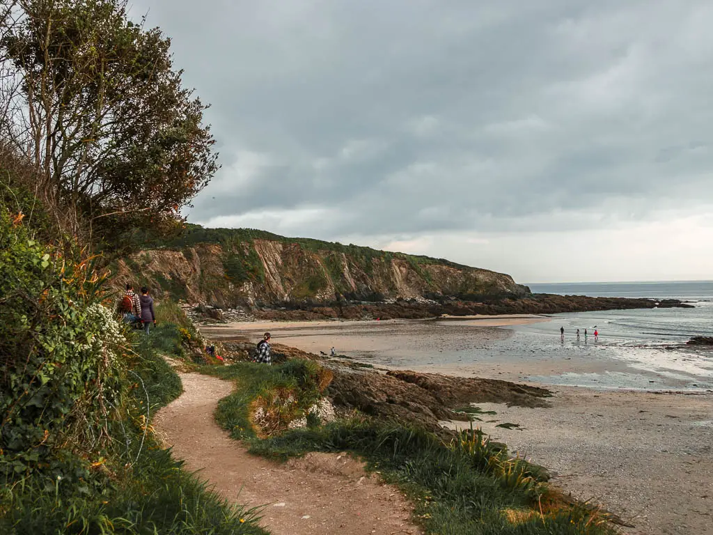 A trail on the left, with the beach and sea to the right, and the cliff face ahead, along the coastal par to Fowey walk. There are a few people on the trail ahead, and some people down on the beach.