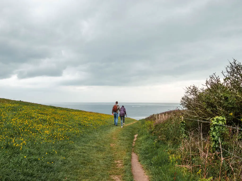 A trail running along a green field with some yellow flowers and a couple of people walking ahead, with a view to the sea beyond that.