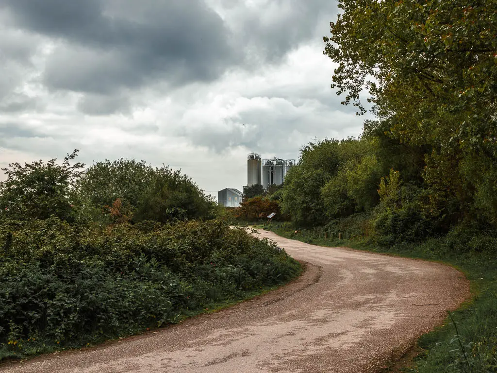 A road curving towards some tall industrial buildings. There are bushes on the left side of the trail, and bushes and trees on the right.