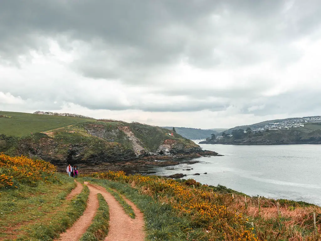 The trail leading down and to the left, with the sea and coastline peninsulars ahead and right near the end of the coastal walk from Park to Fowey. There are a couple of people walking on the trail ahead.